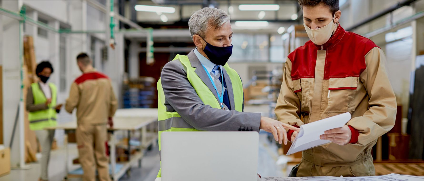Factory worker and businessman with protective face masks analyzing reports at woodworking production facility.